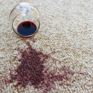 A glass of red wine sitting on a beige carpet with a red wine stain next to it.